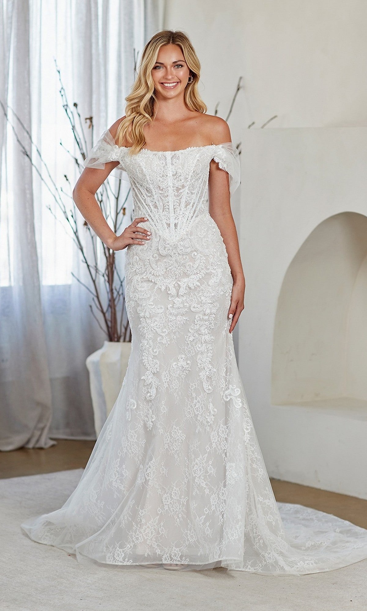A woman smiles at the camera indoors, wearing the Amelia Couture 2153 long prom dress—a fitted off-the-shoulder white lace gown with a flared hem.